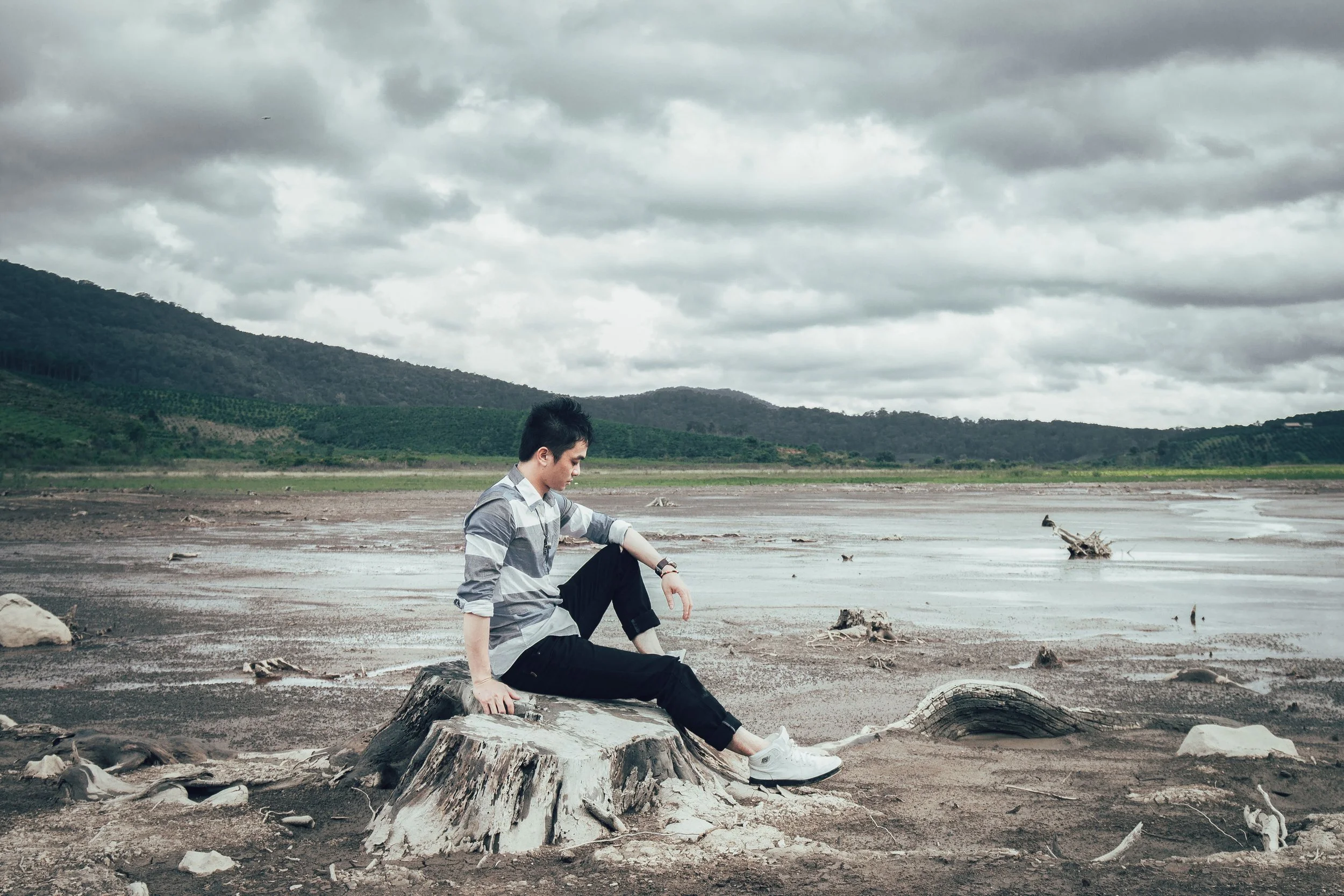 A young man in a striped shirt and black pants sitting on a large tree stump in a muddy, barren landscape with hills and cloudy sky in the background.