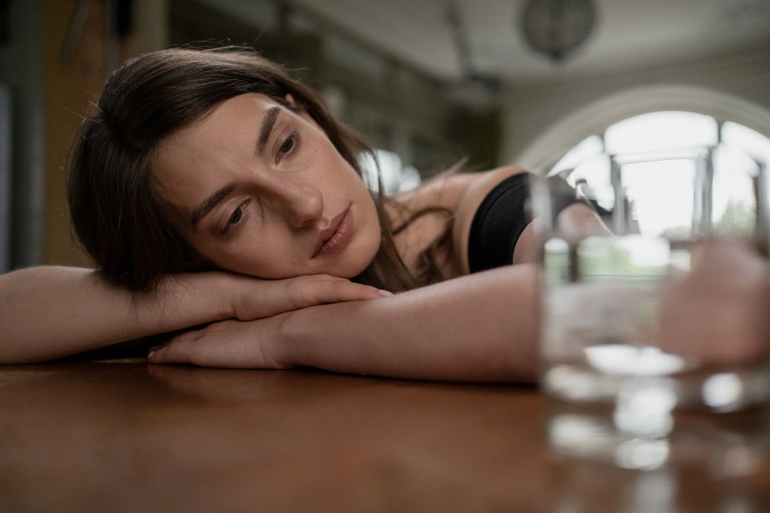 A woman lies resting her head on her arms on a table, looking at bottles of pills in front of her, with a contemplative or concerned expression.