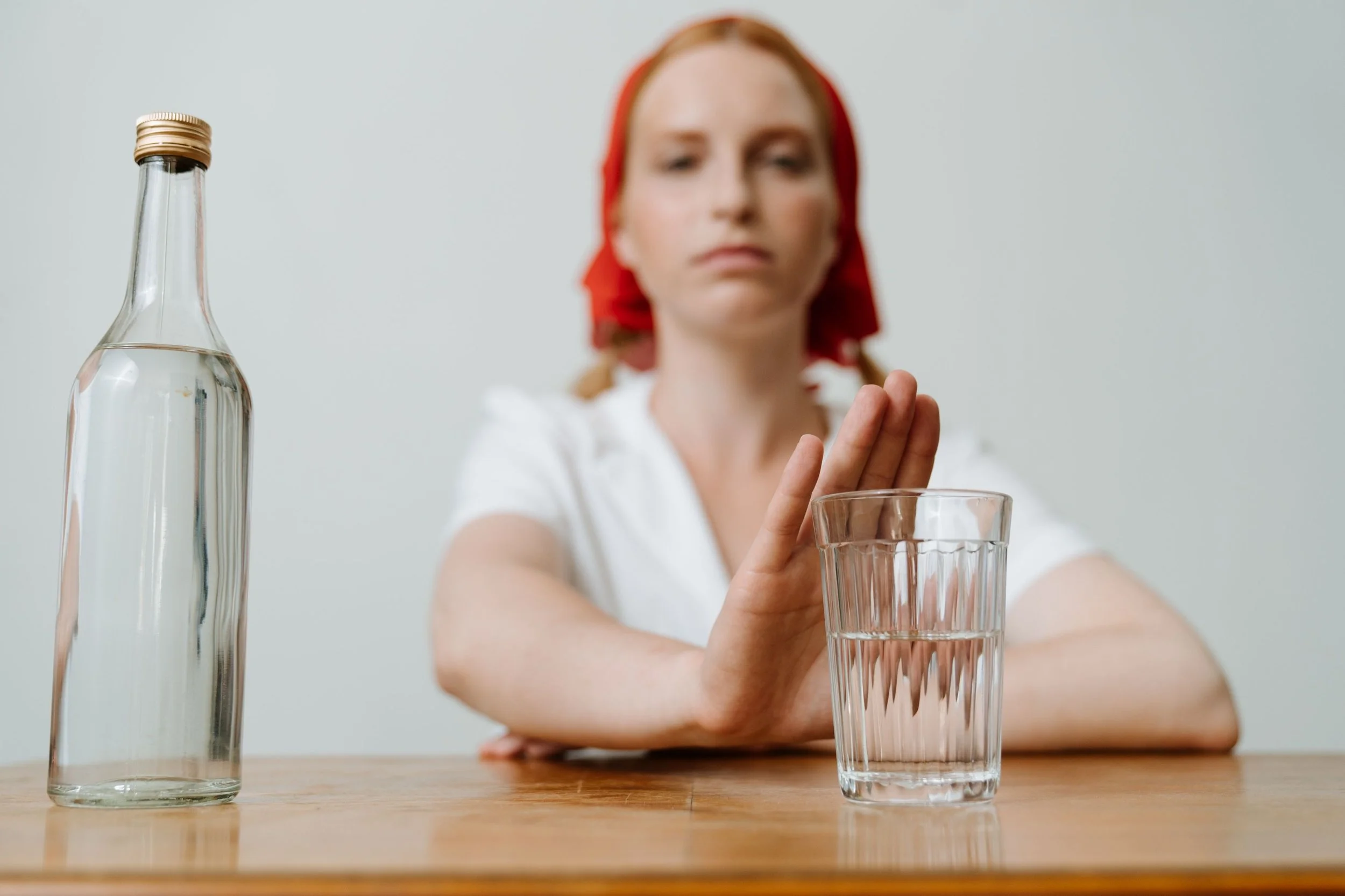 A woman with red hair, wearing a red headscarf, seated at a wooden table, with a clear glass of water in front of her. She is extending her hand towards the glass, and there is a tall, empty glass bottle on the table to her left. The background is plain and light-colored.