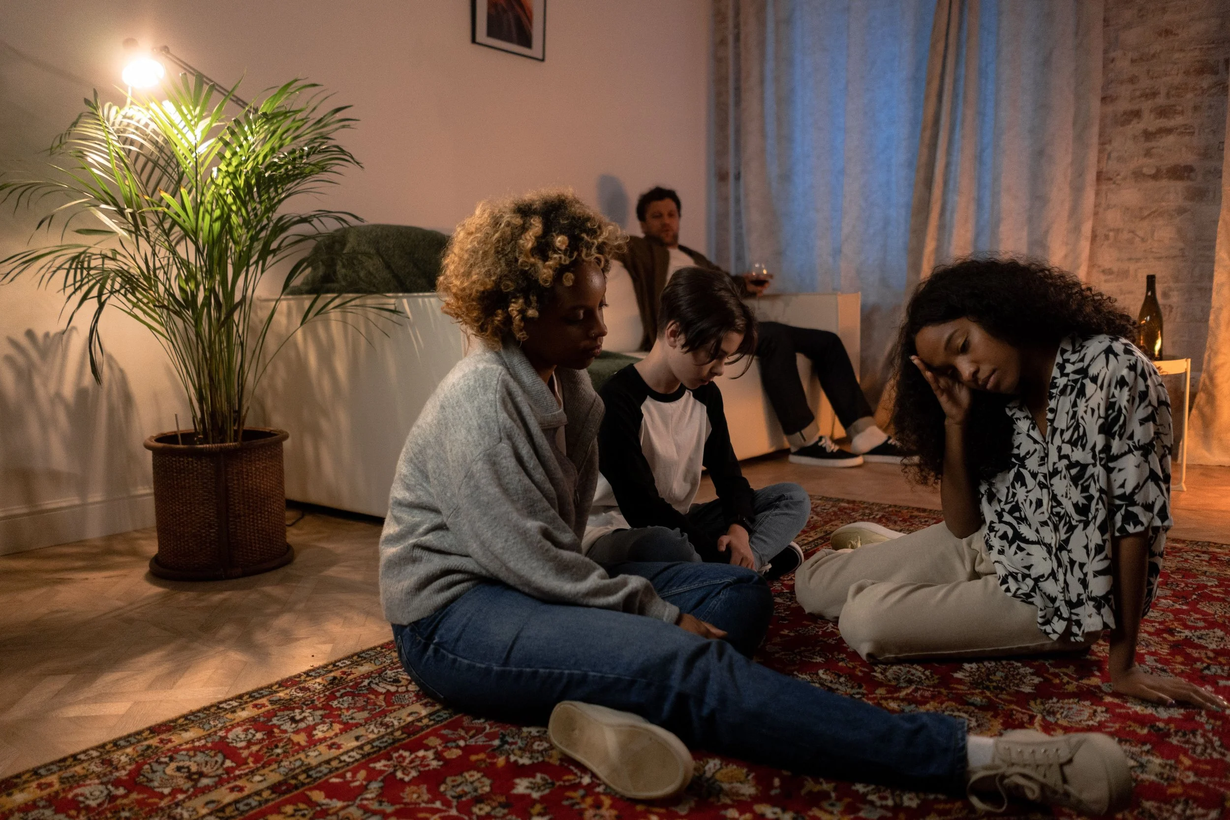 A group of people sitting on a rug in a cozy living room, appearing to be engaged in a shared moment of reflection or prayer, with a potted plant, a lamp, and artwork on the wall.