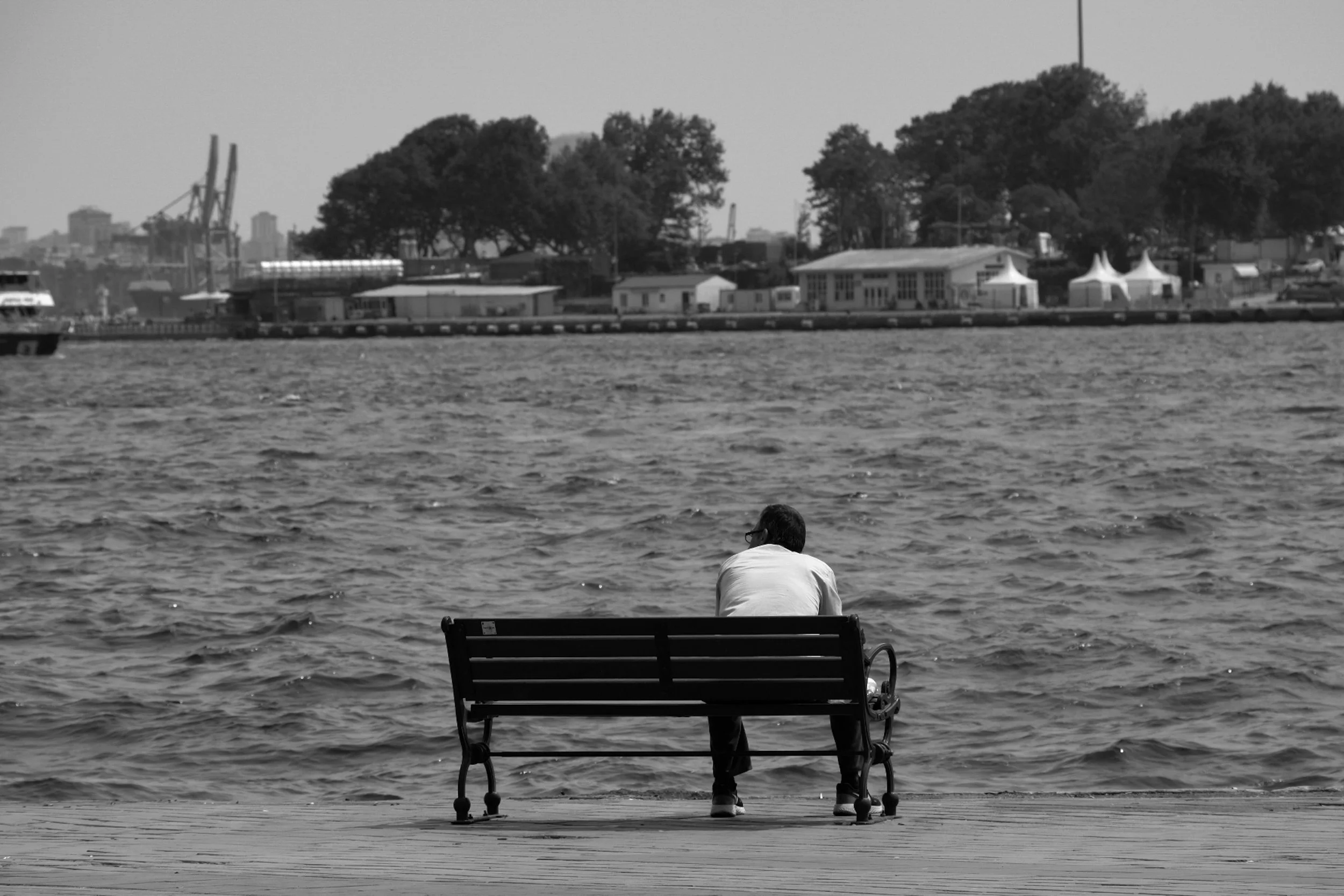 A person sitting alone on a bench near the water, facing away, with trees, buildings, and tents across the river in the background.